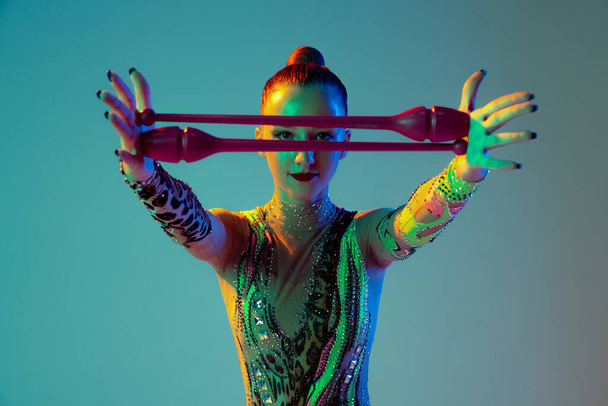 One young rhythmic gymnast posing with clubs in front of her face isolated on gradient green background in neon light. Concept of action, motion, sport life, motivation, competition. Copyspace for ad - Photo, image