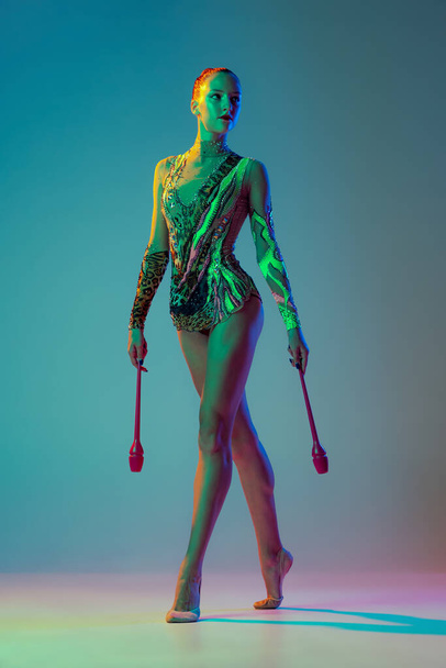 One young sportive girl, female rhythmic gymnast posing with clubs isolated on gradient blue background in neon light. Concept of action, motion, sport life, motivation, competition. Copyspace for ad - Foto, immagini