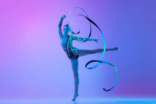 Portrait of one young rhythmic gymnast posing with ribbon in her hand isolated on pink purple background in neon light. Concept of action, motion, sport life, motivation, competition. Copyspace for ad - Photo, image