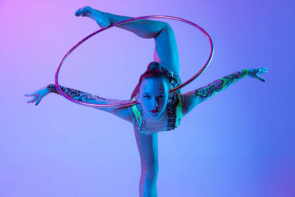 Portrait of young, muscular girl, female rhytmic gymnast training with hoop isolated over blue pink studio background in neon light. Concept of action, motion, sport life, motivation, competition. - Valokuva, kuva