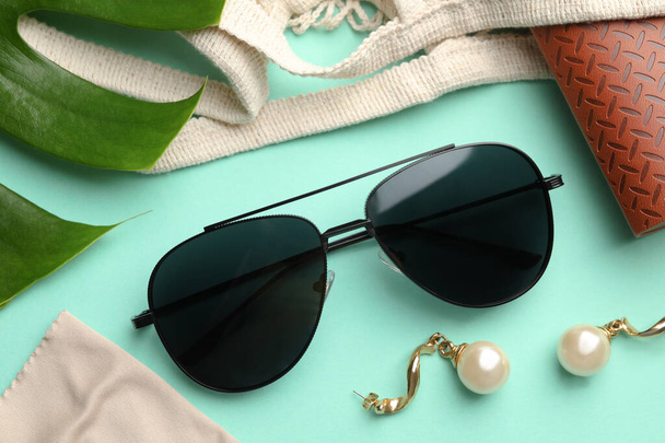 Stylish elegant sunglasses with brown leather case and earrings on turquoise background, flat lay - Foto, Imagem