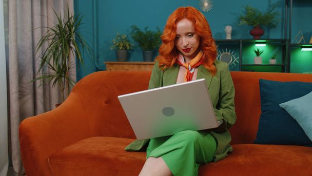 Руда молода жінка в зеленій сукні фрілансер входить до домашньої вітальні і сідає на диван, відкриває ноутбук. Дівчина-імбир працює на ноутбуці, надсилає повідомлення, здійснює онлайн-покупки, переглядає фільми
 - Фото, зображення