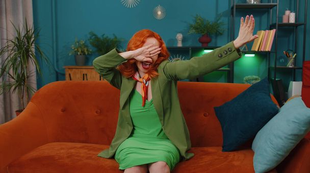Trendy cheerful positive redhead young woman having fun dancing and moving to rhythm, dabbing raising hands, making dub dance gesture at modern home apartment. Ginger girl in living room on sofa - Zdjęcie, obraz