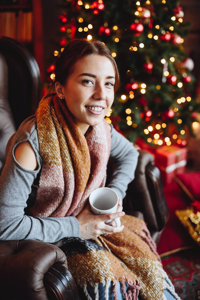Portrait of Smiling Woman Sitting in an Armchair near Christmas Tree with Red Toys with Cup in Her Hands. New Year's Magical New Year's Eve Mood. - Фото, изображение