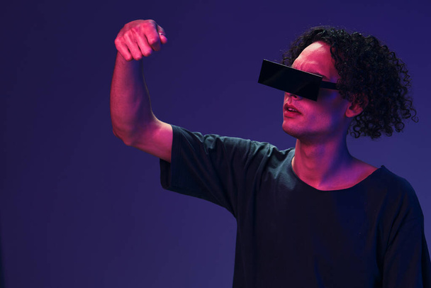 Vzrušený stylový opálený kudrnatý muž v černém tričku s brýlemi drží neviditelný objekt pózující izolovaně na růžovém modrém pozadí. Skvělá módní nabídka. Virtuální realita Nová kolekce koncept. Kopírovat prostor - Fotografie, Obrázek