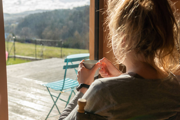 A woman is having morning tea or coffee, relaxing and looking at amazing view at mountains on a bright sunny day. Breathtaking view from a summer house. - Foto, Imagem