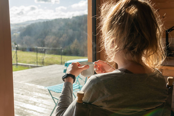 A woman is having morning tea or coffee, relaxing and looking at amazing view at mountains on a bright sunny day. Breathtaking view from a summer house. - Fotografie, Obrázek