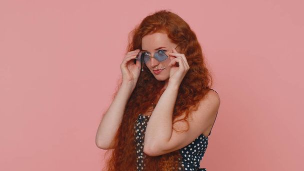 Portrait of seductive cheerful stylish woman in dress wearing sunglasses, charming smile. People emotions concept. Young lovely redhead ginger freckles girl isolated alone on pink studio background - 写真・画像