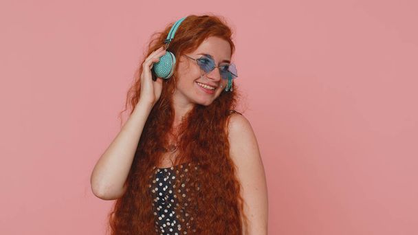 Young redhead woman in dress listening music on headphones, dancing disco fooling around having fun expressive gesticulating hands. Ginger girl with freckles isolated alone on pink studio background - 写真・画像