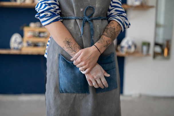 Γυναίκα γλύπτρια καλλιτέχνης φορώντας βρώμικη ποδιά στο στούντιο τέχνης, περικοπεί φωτογραφία. Γυναίκα με τατουάζ κεραμίστρια που στέκεται μέσα σε εργαστήριο κεραμικής, δημιουργώντας και πουλώντας χειροποίητα πήλινα σκεύη, δουλεύοντας με πηλό - Φωτογραφία, εικόνα