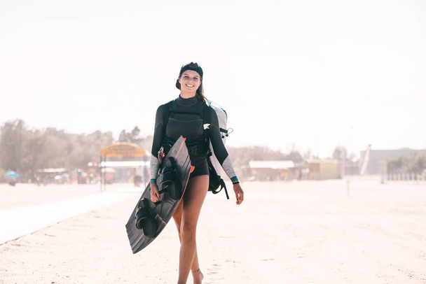 Portrait of beautiful smiling girl posing with a surfboard on the beach and wearing kitesurfing equipment - concept of sporty woman and summer sport activity - Foto, Bild