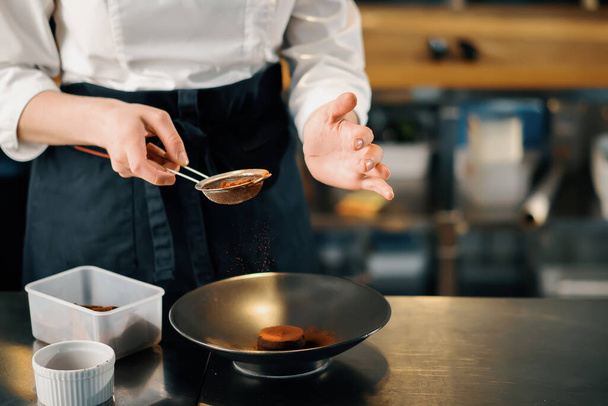 Профессиональная кухня ресторана, крупным планом: женщина-повар посыпает пирожные с шоколадом и корицей - Фото, изображение