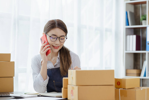 Asian woman is talking on the phone with a customer to confirm an order, she owns an online store, she packs and ships through a private transport company. Online selling and online shopping concepts. - Fotoğraf, Görsel