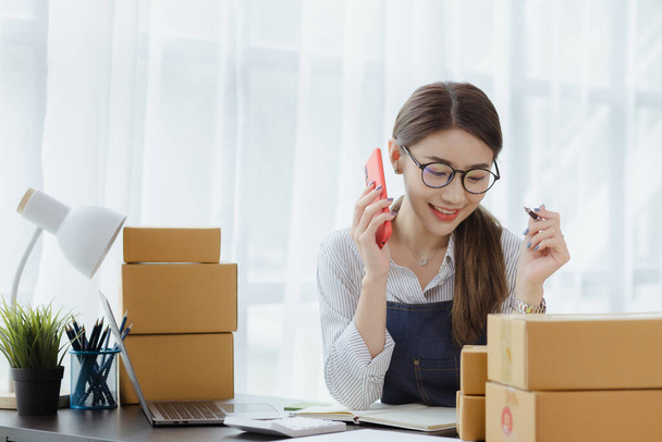 Asian woman is talking on the phone with a customer to confirm an order, she owns an online store, she packs and ships through a private transport company. Online selling and online shopping concepts. - Photo, Image