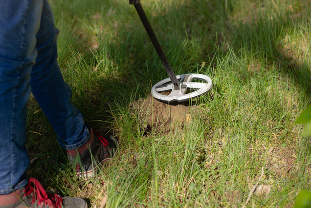 The coil of a wireless metal detector and the legs of a man looking for treasure over a dug hole in the background of the forest. - Foto, afbeelding