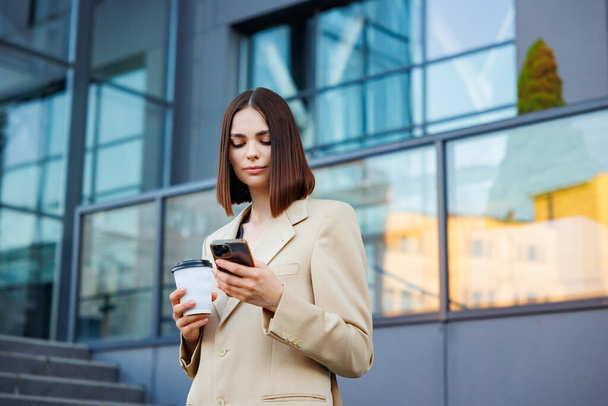 Een jong brunette meisje tegen de achtergrond van een business center, office center. Portret van een succesvolle start-up. Smartphone, koffie casual pak - Foto, afbeelding