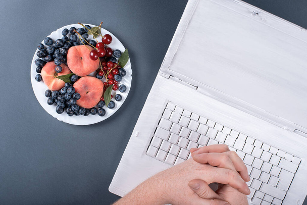 Sommerfrüchte auf grauem Hintergrund auf einem Teller mit einem Laptop in weißer Farbe. Pfirsiche und Kirschen. das Konzept eines Imbisses im Büro für einen Freiberufler - Foto, Bild