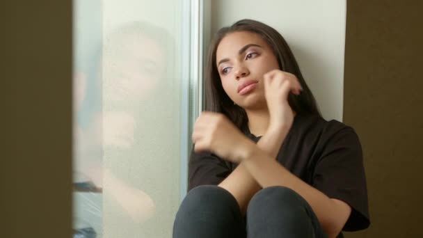 A sad black girl is sitting on the windowsill. A lonely young woman looks thoughtfully out the window. Concept of mental illness, problems. - Footage, Video