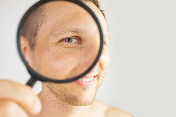 man with magnifying glass near eye mimic wrinkles on face. Portrait isolated on white. Clean healthy skin and cosmetology concept. Image for cosmetology clinic advertising. - Фото, изображение