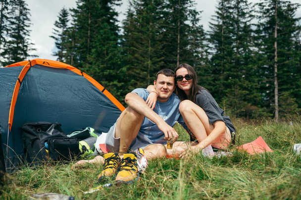 Beautiful young tourist couple man and woman sitting on the grass near a camping site with a tent and looking at the camera. - Fotoğraf, Görsel