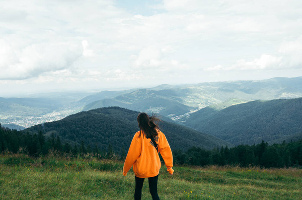 Фото ззаду жінки-туристки в помаранчевій сорочці, що стоїть в горах на лузі і дивиться на красиві краєвиди
 - Фото, зображення