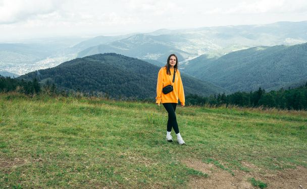 A female tourist in an orange sweatshirt stands in the mountains against the background of beautiful views and poses for the camera with a smile on her face. - Photo, image
