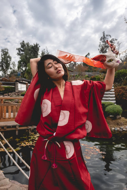 Portrait of sexy and young japanese girl with beautiful old traditional red kimono and handmade cat mask dancing in front of a pond in a japanese garden - Valokuva, kuva