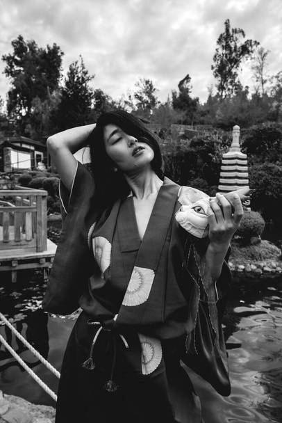 Портрет сексуальной и молодой японской девушки с красивым старым традиционным красным кимоно и кошачьей маской ручной работы, танцующей перед прудом в японском саду (в черно-белом) - Фото, изображение