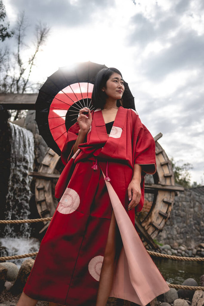 Portrait of sexy and young japanese girl with beautiful old traditional red kimono and handmade red and black paper umbrella in front of a pond with waterfall and mill wheel in a japanese garden - Photo, image