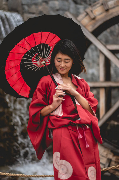 Portrait of sexy and young japanese girl with beautiful old traditional red kimono and handmade red and black paper umbrella in front of a pond with waterfall and mill wheel in a japanese garden - Photo, Image