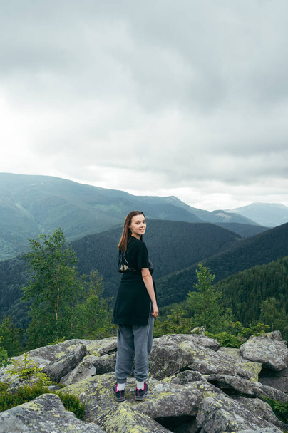 Positive woman in casual clothes stands on a rock in the mountains during a hike and poses for the camera against the background of Carpathian views with a smile on her face. Vertical - Foto, immagini