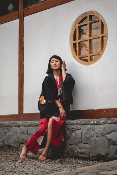 Портрет сексуальної і молодої япанської дівчини з красивими старими традиційними червоними кімоно і чорними юкатами перед старою білою стіною в садку япан. - Фото, зображення