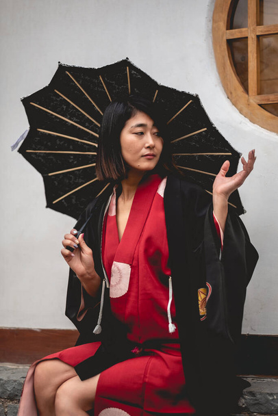 Portrait of sexy and young japanese girl with beautiful old traditional red kimono, black yukata and black lace umbrella in front of a old white wall in a japanese garden - Fotó, kép