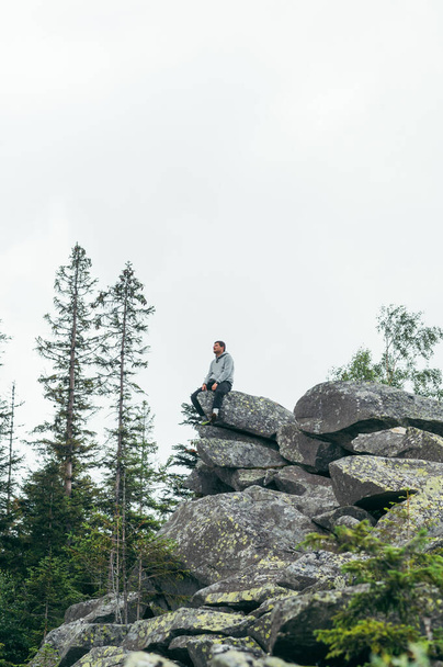 A male extreme tourist sits on the rock of the top of the mountain with his legs hanging down and looks at the landscape of the Carpathians - Photo, Image
