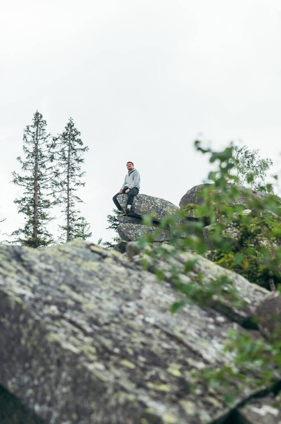 A male tourist in casual clothes sits on a rock on top of a mountain and looks at the views. Active recreation hiking in the mountains. - Фото, зображення