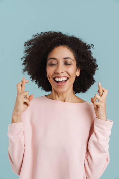 Black young woman smiling while holding fingers crossed for good luck isolated over blue background - Foto, Bild