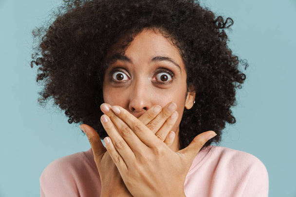Young black woman covering her mouth while expressing surprise isolated over blue background - Fotó, kép