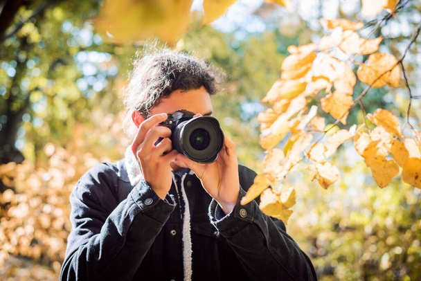 Молодий чоловік фотограф з темним кучерявим волоссям в осінньому парку. Чудова сонячна погода, яскраво освітлені рослини оточують людину
. - Фото, зображення