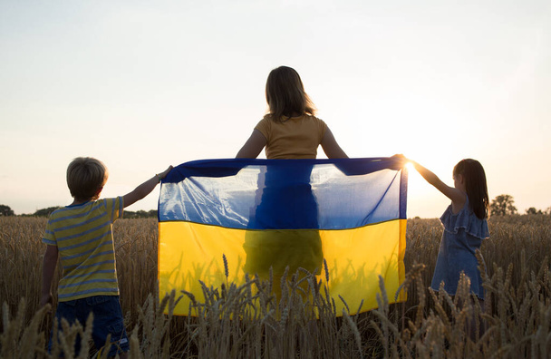 children and a woman stand with their backs against sky, setting sun, wheat field with a blue and yellow Ukrainian flag. Family, refugees, unity, support, patriotism. Ukrainians are against war - Foto, Bild