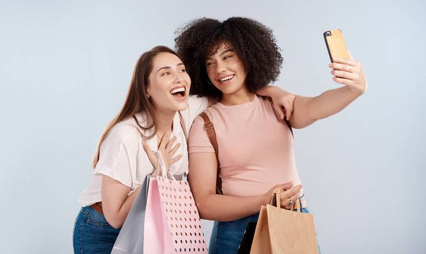 Bargainbuddies. Studio shot of two young women carrying a bunch of shopping bags and taking a selfie - Photo, Image