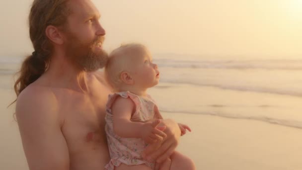 Caucasian bearded father holding little baby infant small daughter son newborn standing at sandy beach near sea ocean watching in gold sky dad talking explain showing direction with finger pointing - Filmati, video