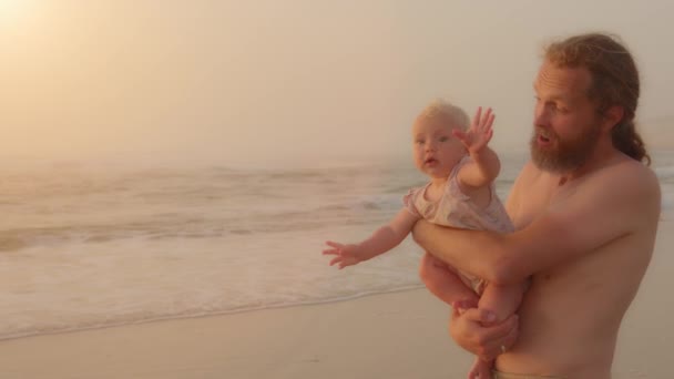 Caucasian smiling family father carrying holding little baby infant small daughter son greeting hello gesture greet looking at camera standing at tropical sandy beach near sea water seaside vacation - Séquence, vidéo
