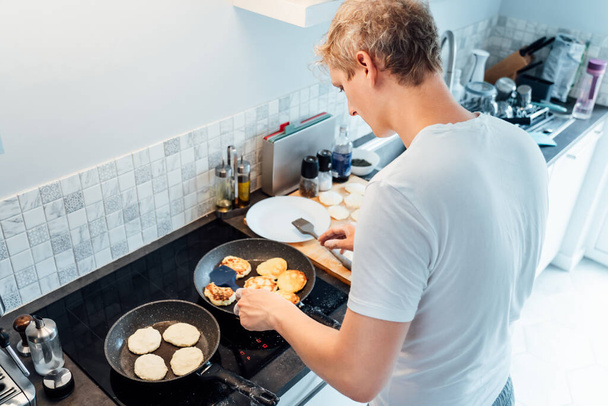 A young man is cooking cheese pancakes in a modern light kitchen at home. Man preparing pancakes for frying on the pan. Housework and home duties. Culinary hobby.High view. Selective focus - 写真・画像