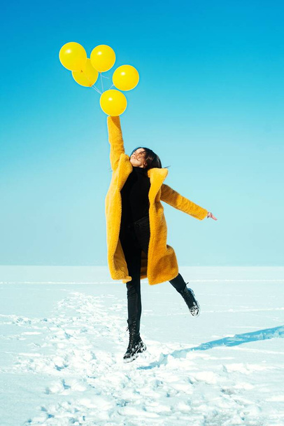 Mujer joven corriendo con abrigo amarillo caminando sobre hielo nevado. con globos amarillos - Foto, imagen