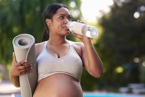 Η ενυδάτωση είναι απαραίτητη, ειδικά όταν είσαι έγκυος. έγκυος γυναίκα που κρατάει το στρώμα γιόγκα και πίνει νερό ενώ στέκεται έξω - Φωτογραφία, εικόνα