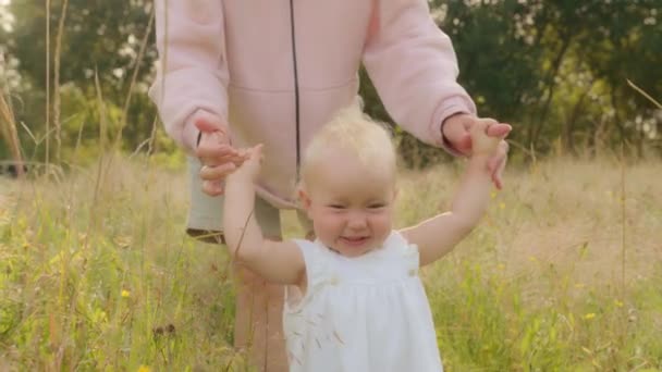 Happy toddler smiling walks through meadow in green high grass holding unrecognizable mother by hand. Mom holding kid daughter baby helps teaches walking in park. First steps of little baby outdoors - Séquence, vidéo