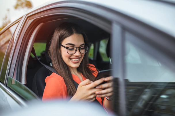 Beautiful business woman is using a smart phone and smiling while sitting on front seat in the car. Portrait of beautiful smiling happy woman in a car. Beautiful business woman in car. - Фото, изображение