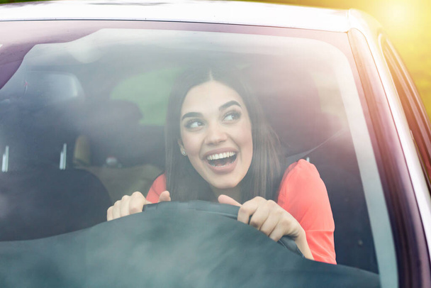 Happy brunette woman driving a car. Portrait of beautiful caucasian woman with toothy smile and brown hair driving car. Hand on steering wheel. Young woman driving a car in the city - Fotó, kép