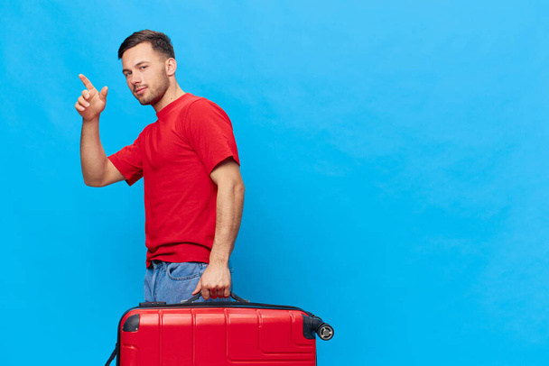 Ein fröhlicher junger, braun gebrannter, gutaussehender Mann in rotem T-Shirt, bereit für den Urlaub, winkt zum Abschied mit einem Koffer und posiert isoliert auf blauem Studiohintergrund. Kopierraum Banner-Attrappe. Reisekonzept - Foto, Bild