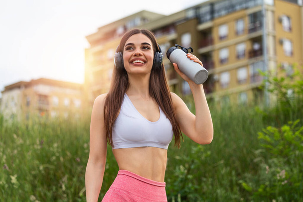 Urban sport girl with headphones drinking water in sunset. excited happy young woman smiling to sun with bottle of water. Attractive sportswoman, enjoying summer, training, outwork, happiness - Фото, изображение